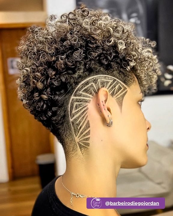Voluminous curly frohawk featuring ash blonde highlights on top, paired with low-faded sides and a sharp geometric shaved undercut pattern.