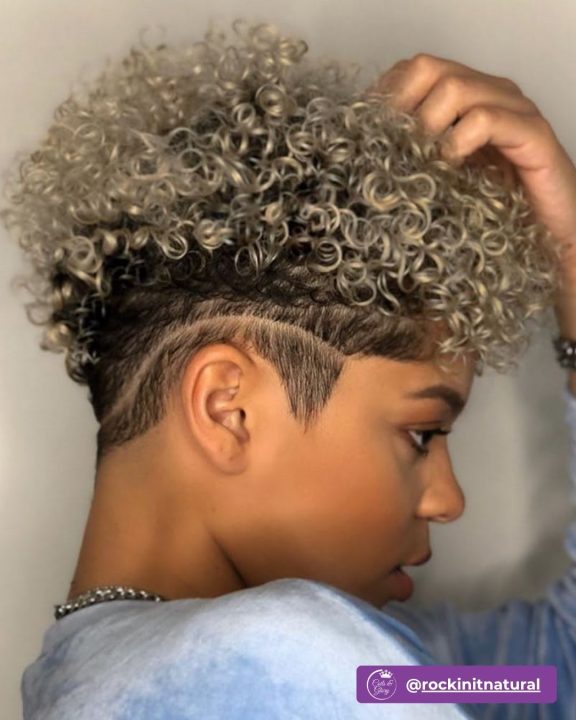 Close-up of a high-volume curly haircut with ash blonde tones on top, neat tapered sides, and a single precise razor slit shaved into the temple.