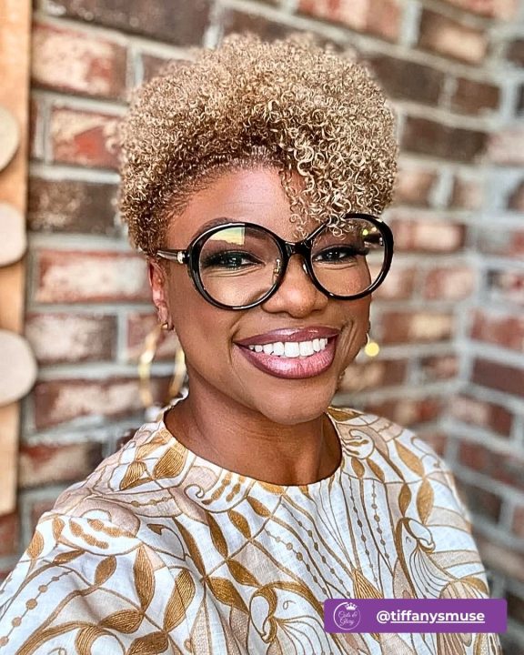 Warm honey blonde coiled haircut tapered closely along the sides and back, styled with soft ringlet curls falling forward into a chic fringe.