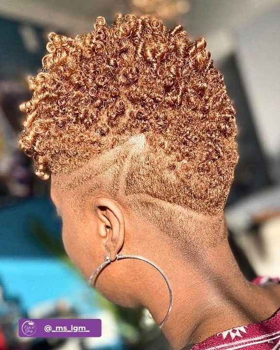 Warm caramel blonde coiled curls tapered along the sides and back, paired with a sleek fade carved with a precise geometric razor pattern.