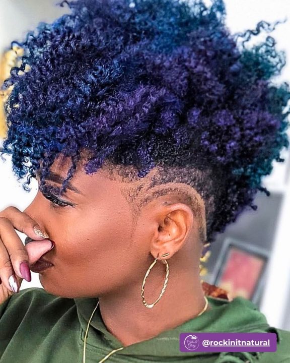 Vibrant indigo-violet curly afro tapered along the sides and nape, accented with a sleek, curved wave line carved into the temple fade for a bold women's short natural look.