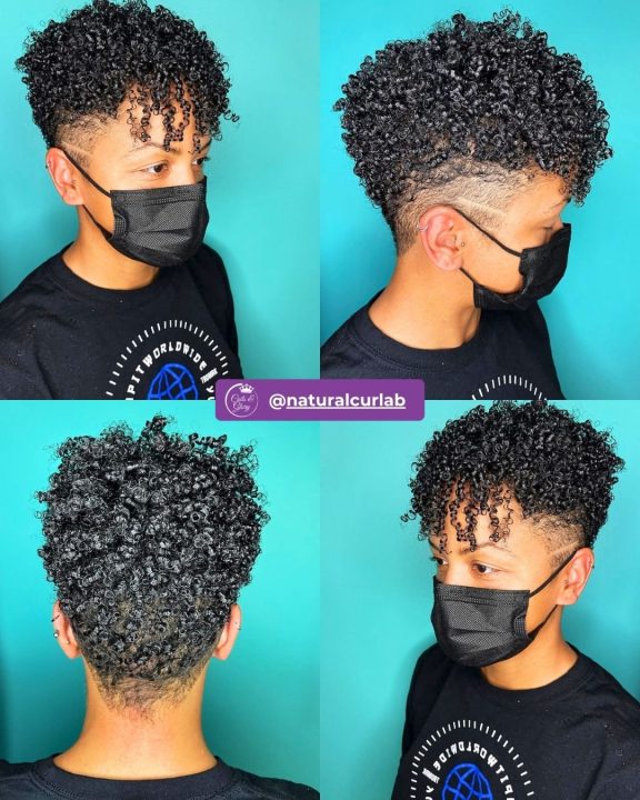 Deep jet black curly pixie crop with defined natural texture on top, paired with neatly tapered sides accented by a sharp, single razor side slit line.