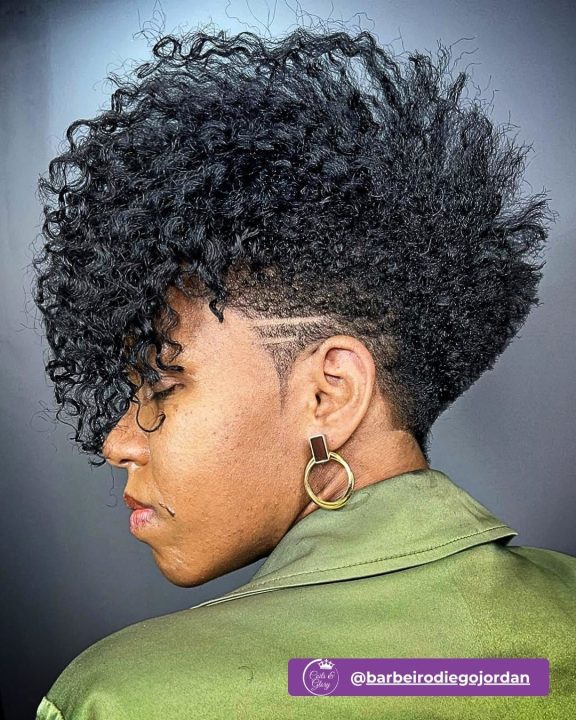 Rich natural black curly frohawk with defined volume running along the center, contrasted by neatly tapered sides accented with sharp razor side slits.