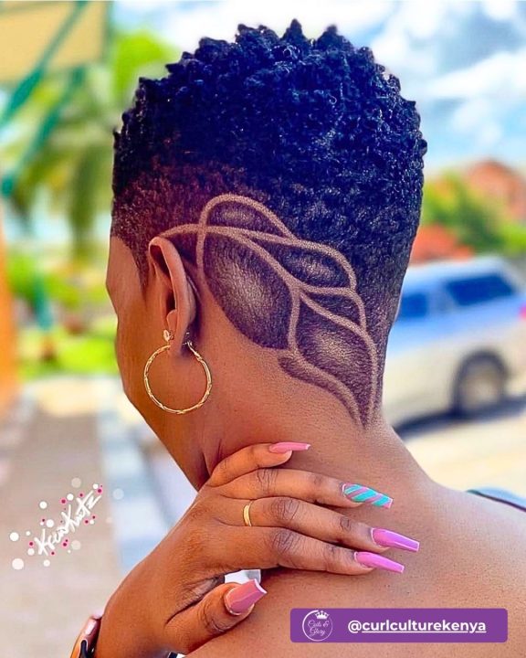 Rich natural black short afro with defined texture on top, paired with a clean taper fade featuring a detailed leaf pattern carved into the undercut along the side and nape.